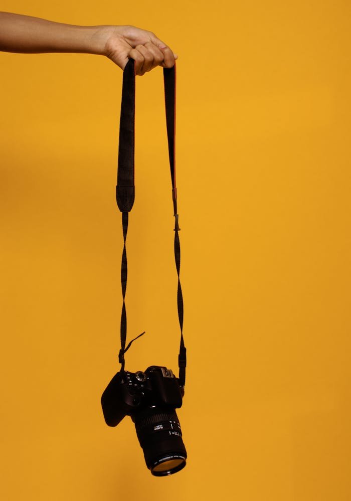 A DSLR camera suspended by hand against a vibrant orange backdrop, showcasing photography technology.